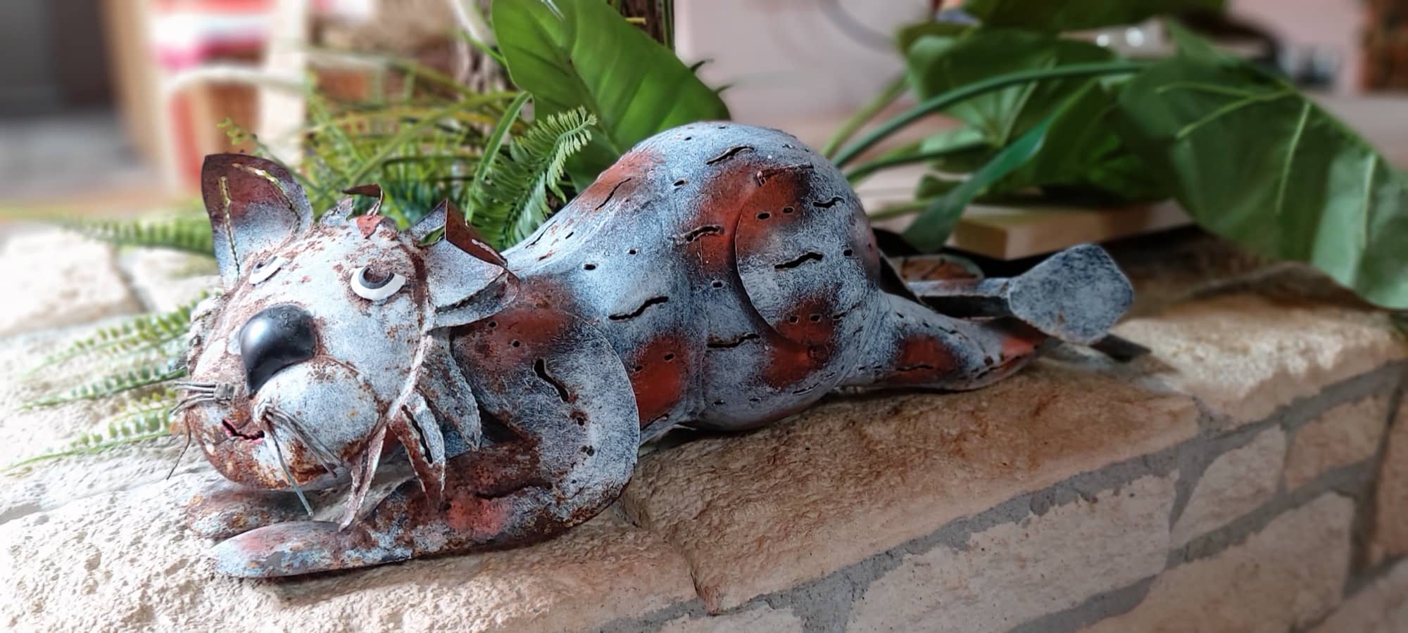Katzenfigur aus Metall auf einer Sandsteinmauer zwischen Grünpflanzen